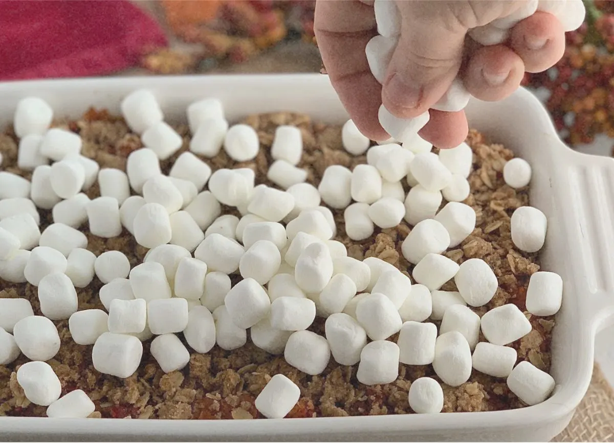 A hand sprinkling some marshmallows on a gluten and dairy free sweet potato casserole in a baking dish.