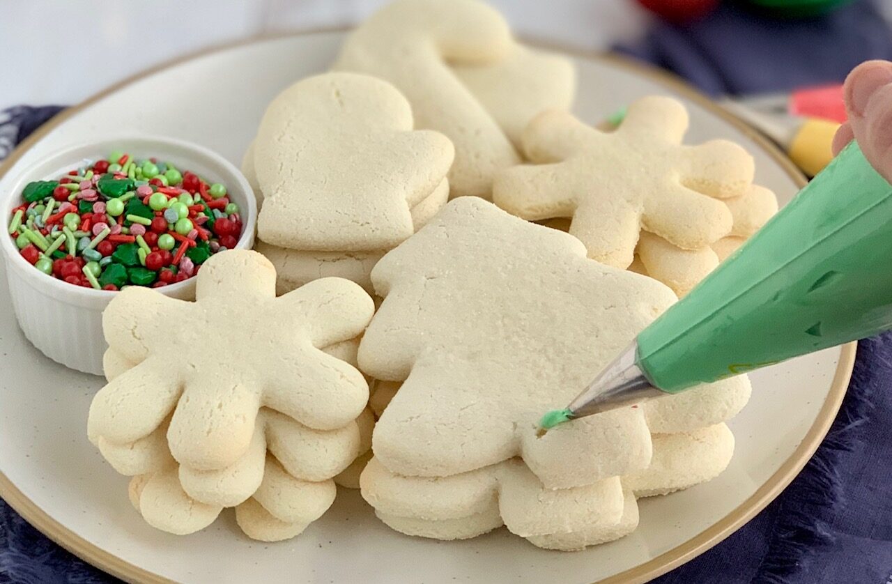 A plate with several baked white cookies in different shapes. Some are Christmas tr3ees, candy canes, mittens, and snowflakes with a little bowl of festive sprinkles..