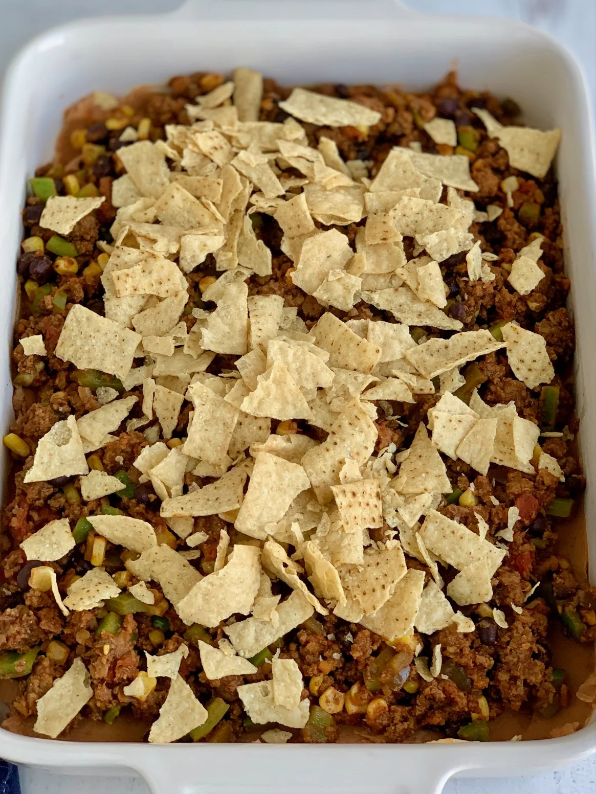 A baking dish filled with a layer of refried beans and taco meat sauce and crunched tortilla chips.