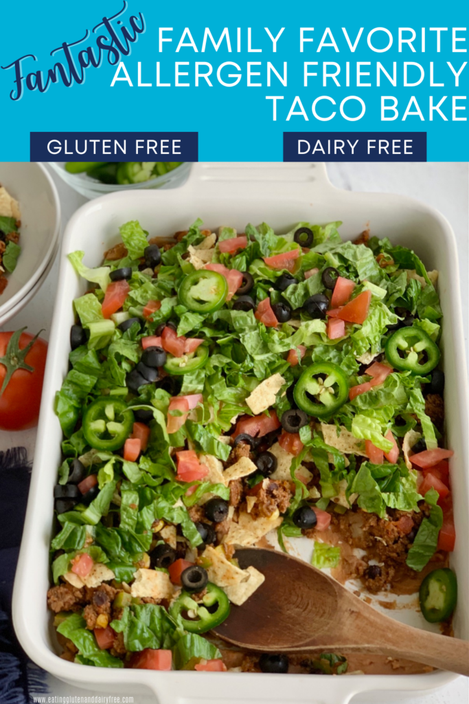 A baking dish filled with a layer of refried beans, taco meat sauce, crunched up chips, shredded romaine lettuce, diced tomatoes, jalapeno peppers, and black olives. Several scoops have been taken out.