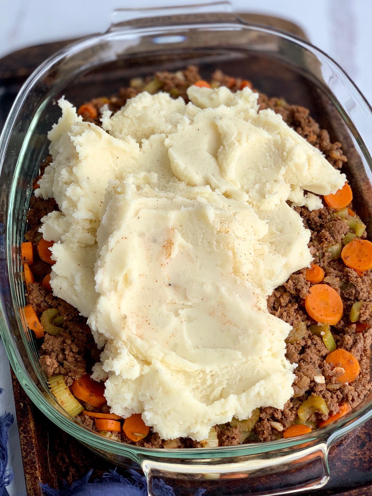 A 9x13-inch glass baking pan with a seasoned ground beef, carrot, and celery mixture topped with creamy, thick mashed potatoes before it goes into the oven.