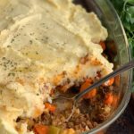 A 9x13-inch glass baking pan with a seasoned ground beef, carrot, and celery mixture topped with creamy, thick mashed potatoes. Melted dairy free butter and little fresh parsley specks are on top of the mashed potatoes. A spoon has scooped out some of the shepherd's pie.