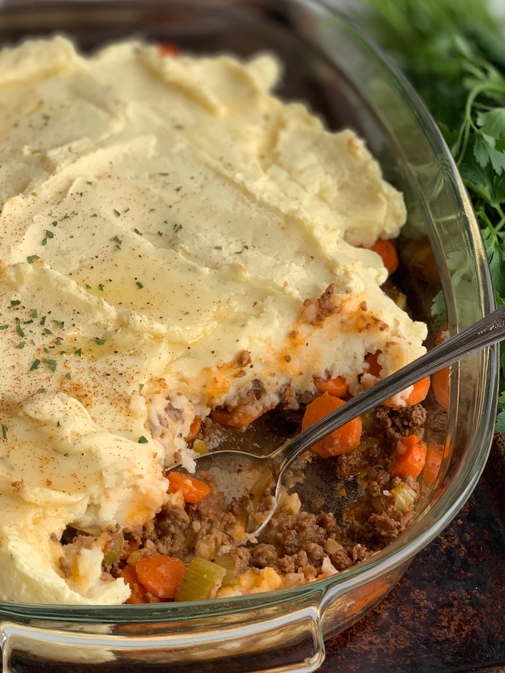 A 9x13-inch glass baking pan with a seasoned ground beef, carrot, and celery mixture topped with creamy, thick mashed potatoes. Melted dairy free butter and little fresh parsley specks are on top of the mashed potatoes. A spoon has scooped out some of the shepherd's pie. 