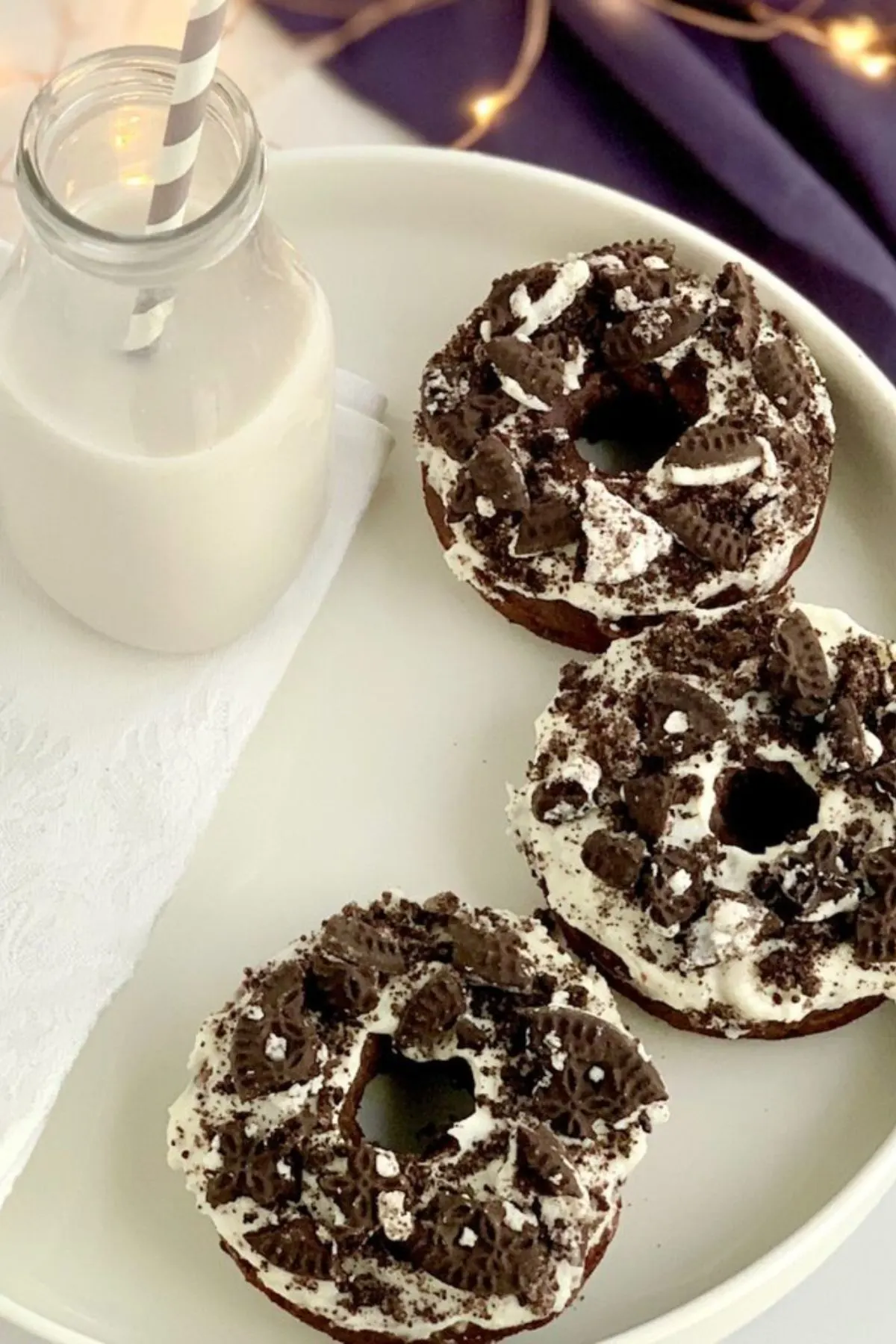3 oreo donuts with frosting and crushed oreo cookies on top next to a glass of milk. 