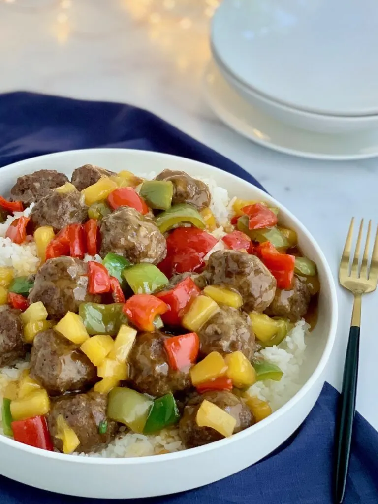 A white serving platter of steamed white rice with beef meatballs, pineapple chunks, and red, green and orange bell peppers.