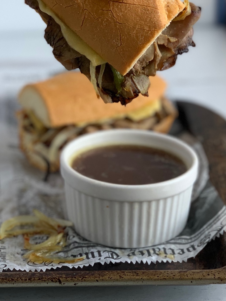 A sliced baguette with sautÃ©ed onions and green peppers and roast beef being dipped into a Au Jus.