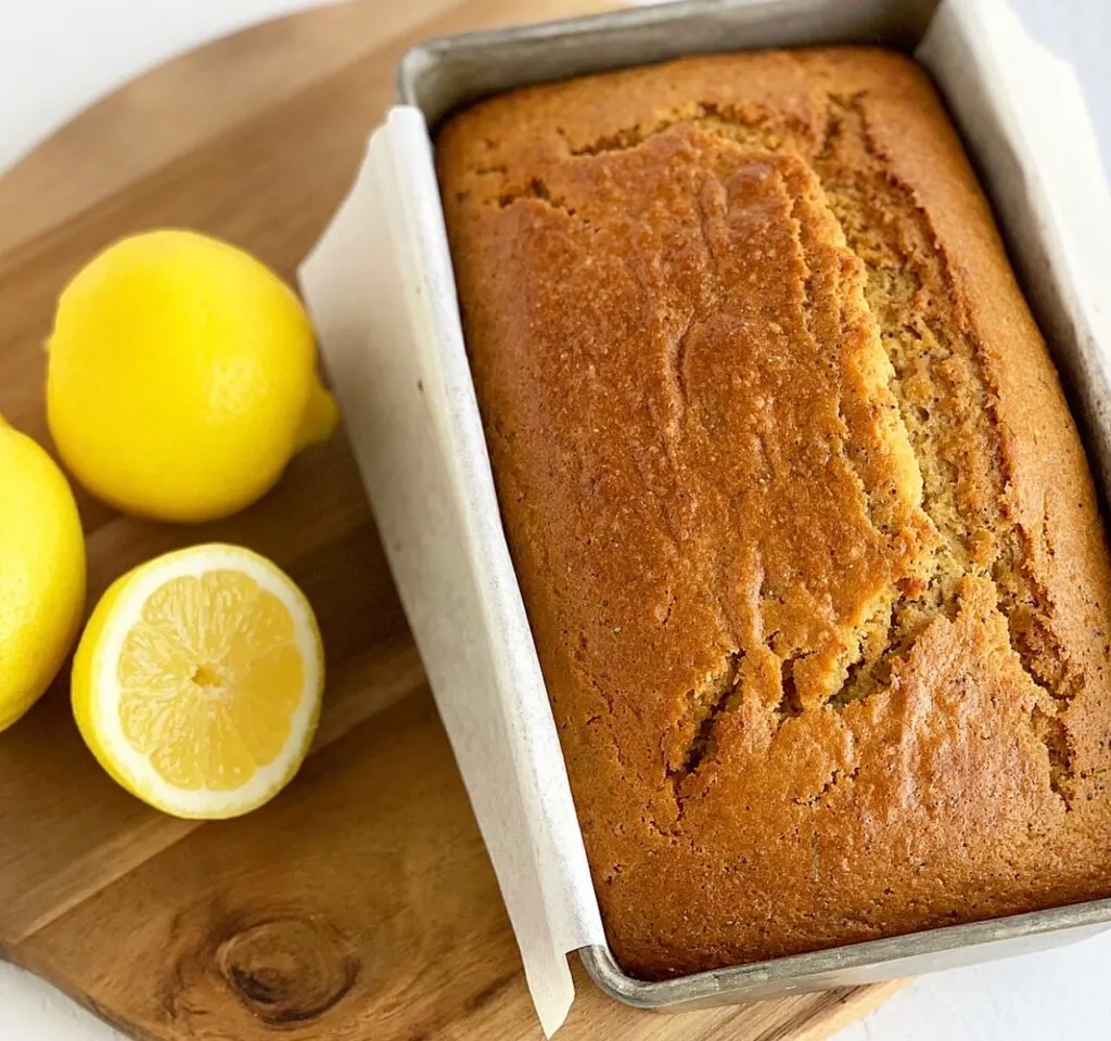 A baked loaf of Lemon Poppy Seed Bread in loaf pan next to three lemons.