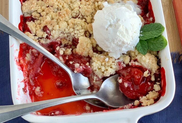 A white baking dish filled with slices of strawberries and rhubarb in a thick juice topped with a buttery flour mixture. Two spoons are in the dish next to a dollop of ice cream and mint leaves.