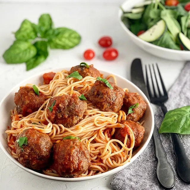 A white bowl with thin spaghetti noodles with red sauce and several baked meatballs on the top next to black silverware.