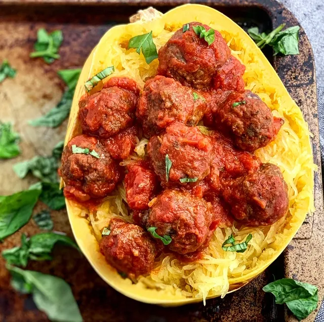 A baked yellow spaghetti squash with several brown meatballs and red spaghettis sauce over the top on a dark baking pan.