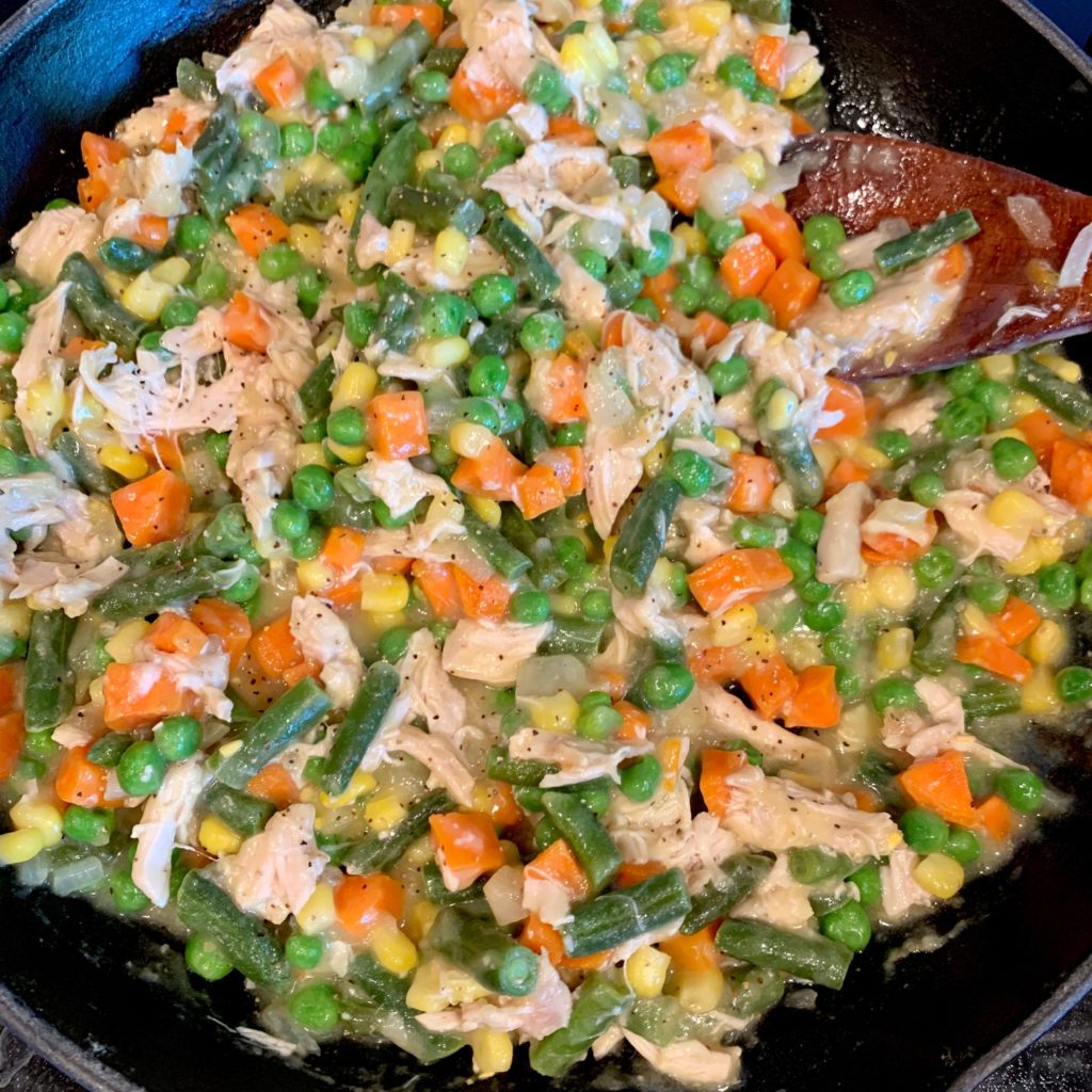 The thick chicken pot pie filling in a skillet with onions, chicken, carrots, corn, peas, and green beans.