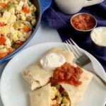 A white plate with 2 large breakfast burritos on it. One is cut open and you can see eggs, bell peppers, sausage, and hash browns. They are topped with dairy free sour cream and a dollop of salsa.