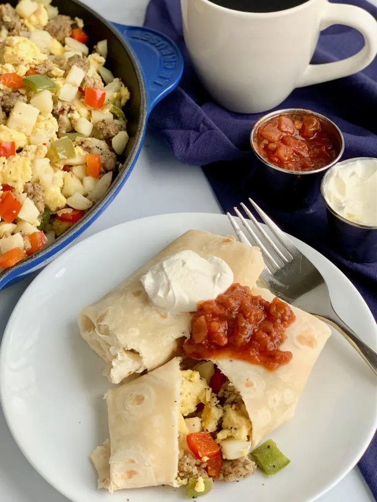 A white plate with 2 large breakfast burritos on it. One is cut open and you can see eggs, bell peppers, sausage, and hash browns. They are topped with dairy free sour cream and a dollop of salsa