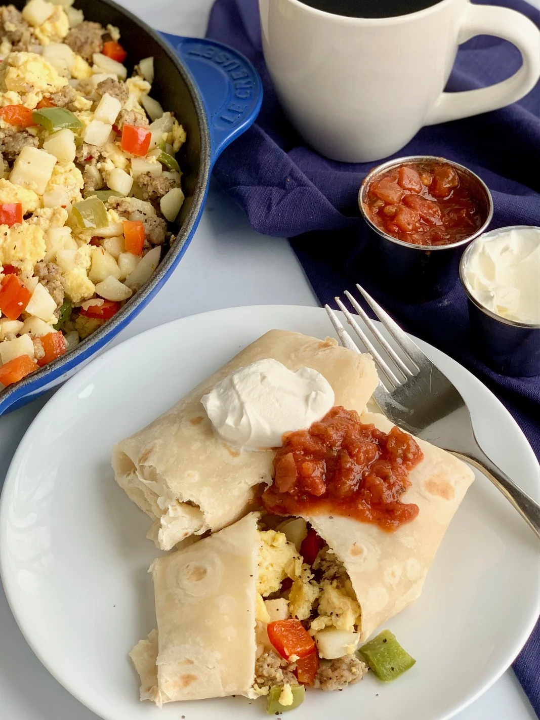 A white plate with 2 large breakfast burritos on it. One is cut open and you can see eggs, bell peppers, sausage, and hash browns. They are topped with dairy free sour cream and a dollop of salsa.