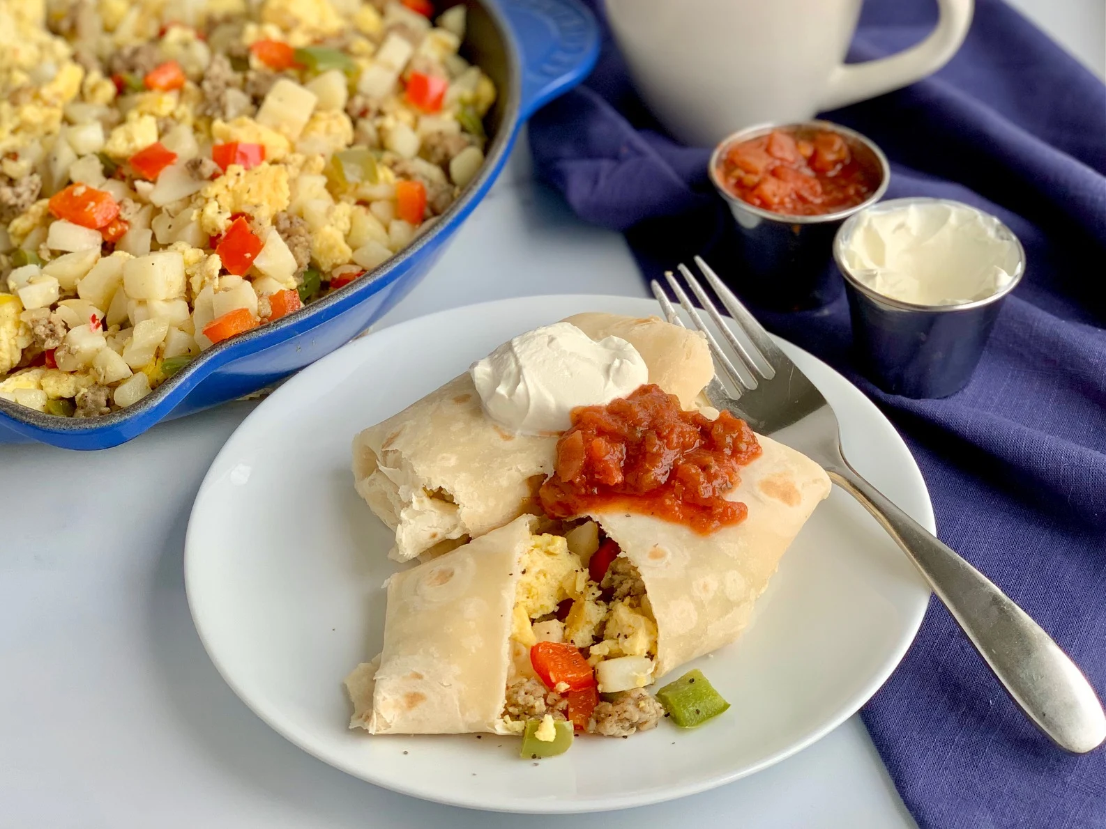 A white plate with 2 large breakfast burritos on it. One is cut open and you can see eggs, bell peppers, sausage, and hash browns. They are topped with dairy free sour cream and a dollop of salsa