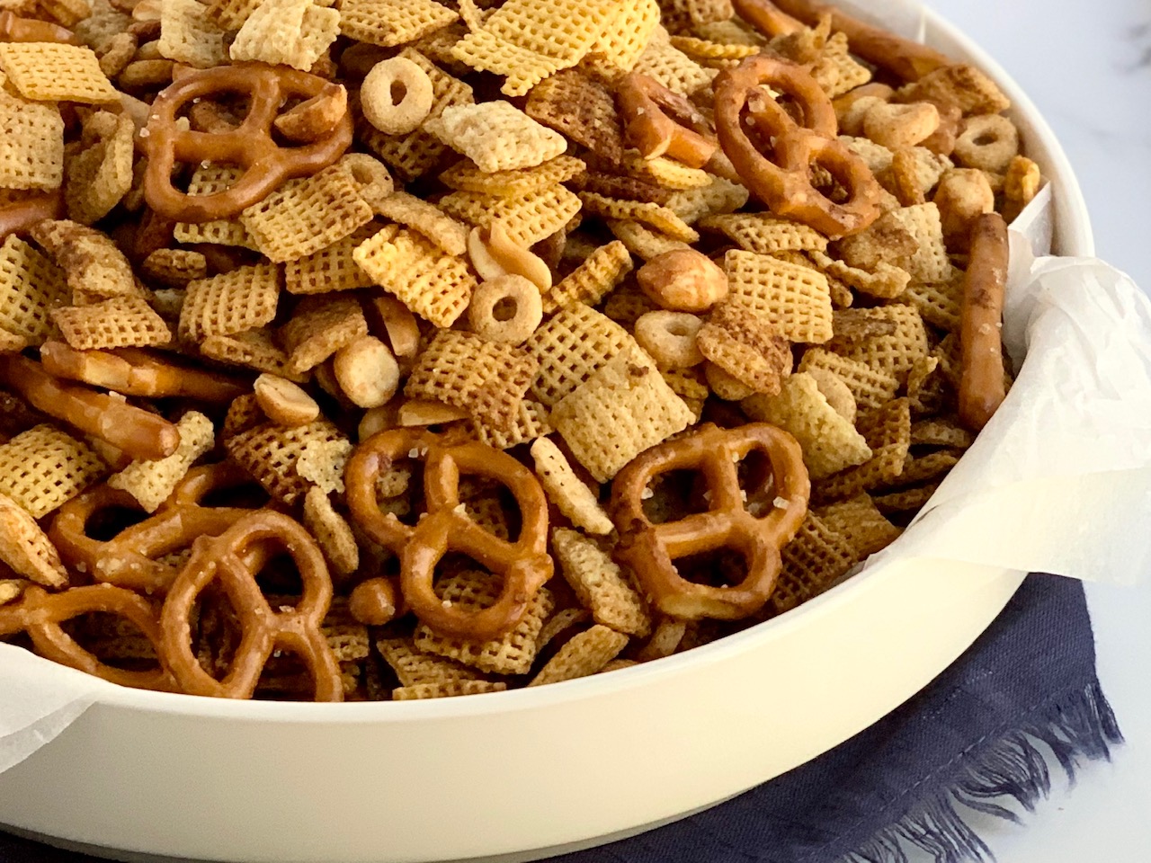 A white platter bowl full of pretzel rods, twists, peanuts, Chex cereal, seasonings, and Whole O's cereal.