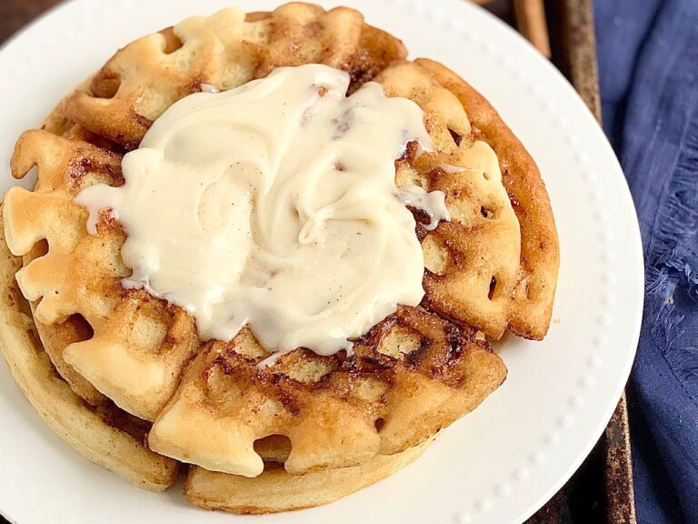 A white plate with 2 large waffles on it. The waffles have a cinnamon swirl mixture in the them and topped with a creamy frosting.
