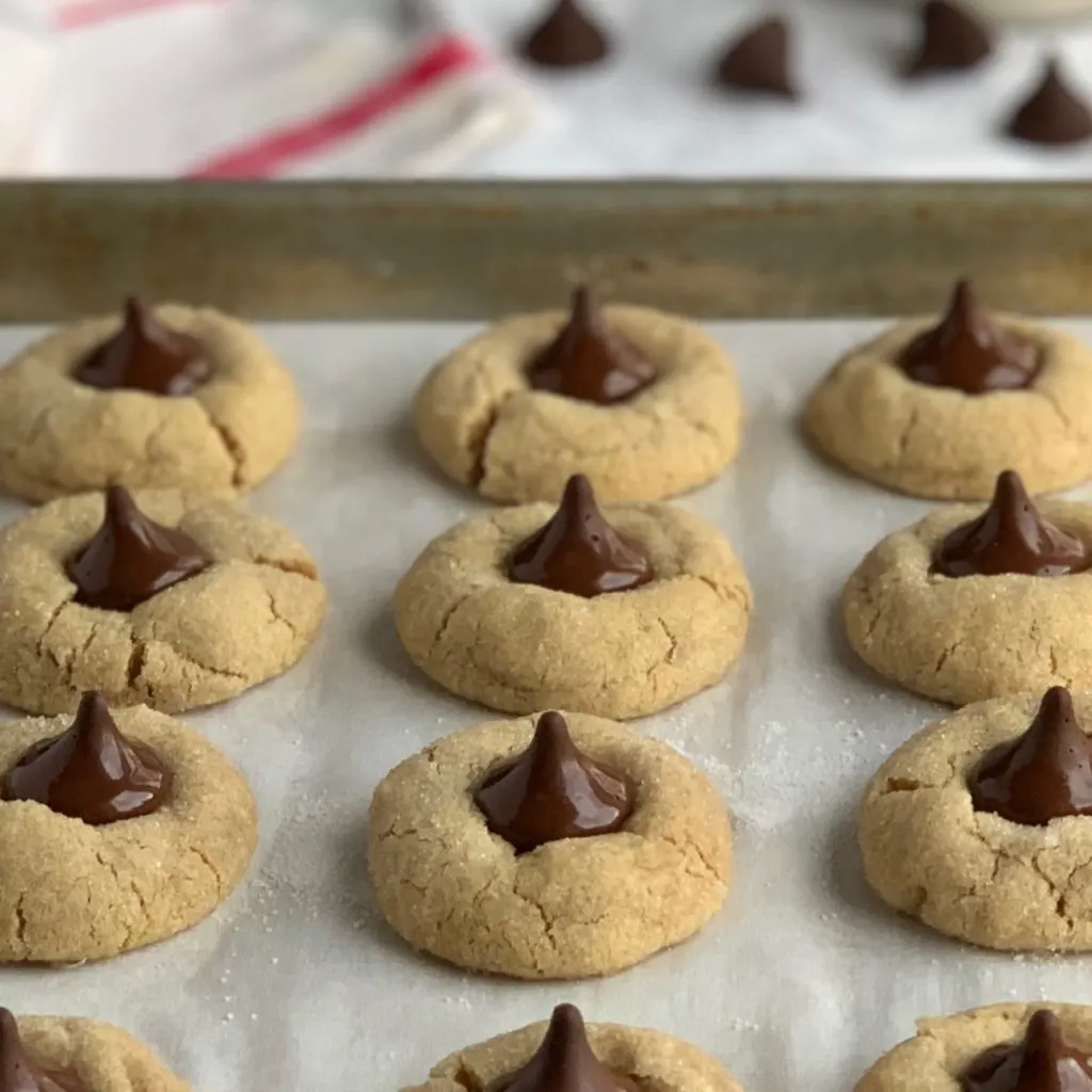 A baking sheet pan full of peanut butter sugar cookies with soft chocolate kisses on them