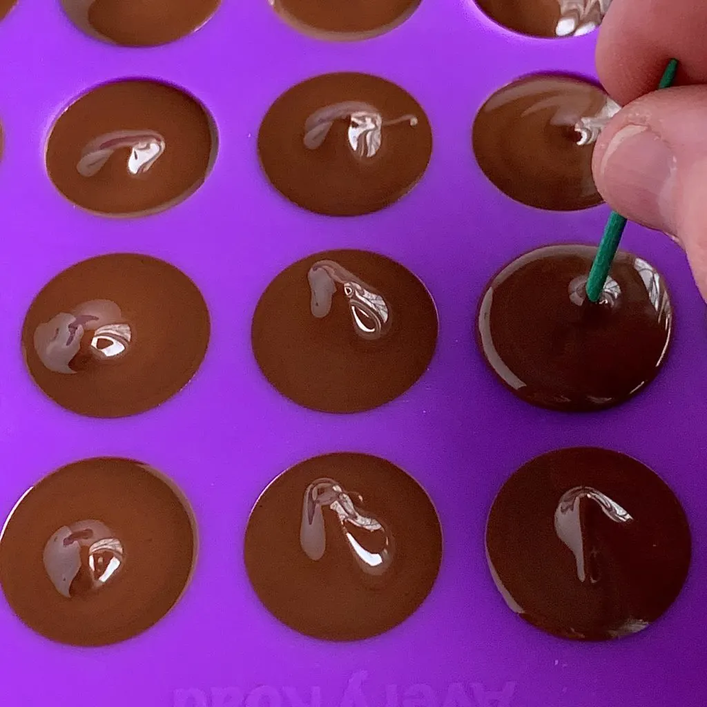 a toothpick pushing melted chocolate into the tip of a chocolate kiss cavity mold.