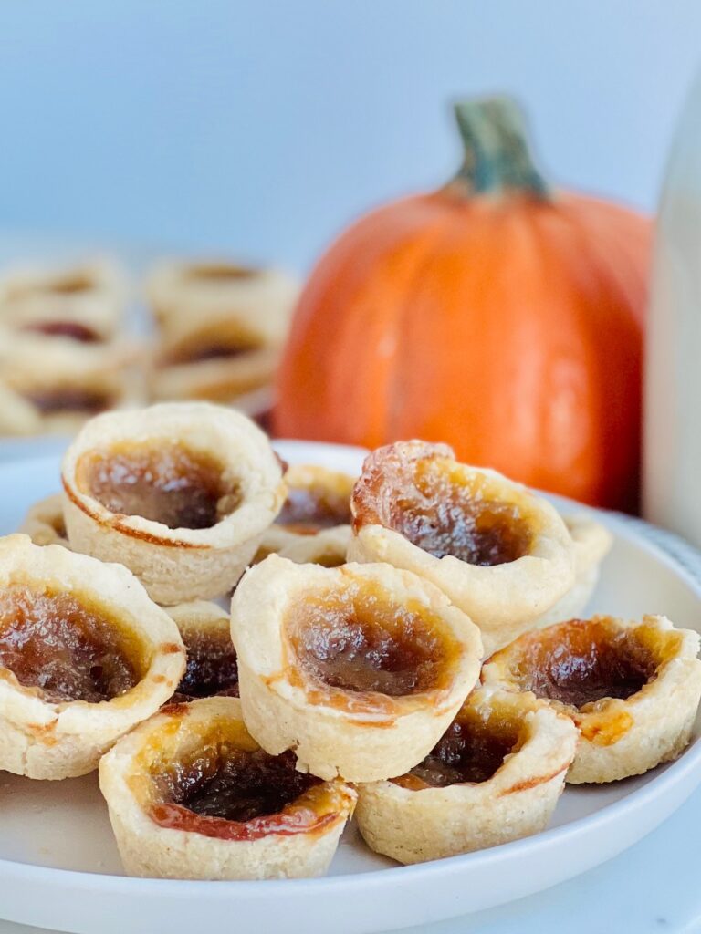 A white plate full of mini little baked tarts filled with a brown sugar gooey mixture with a pumpkin in the background.