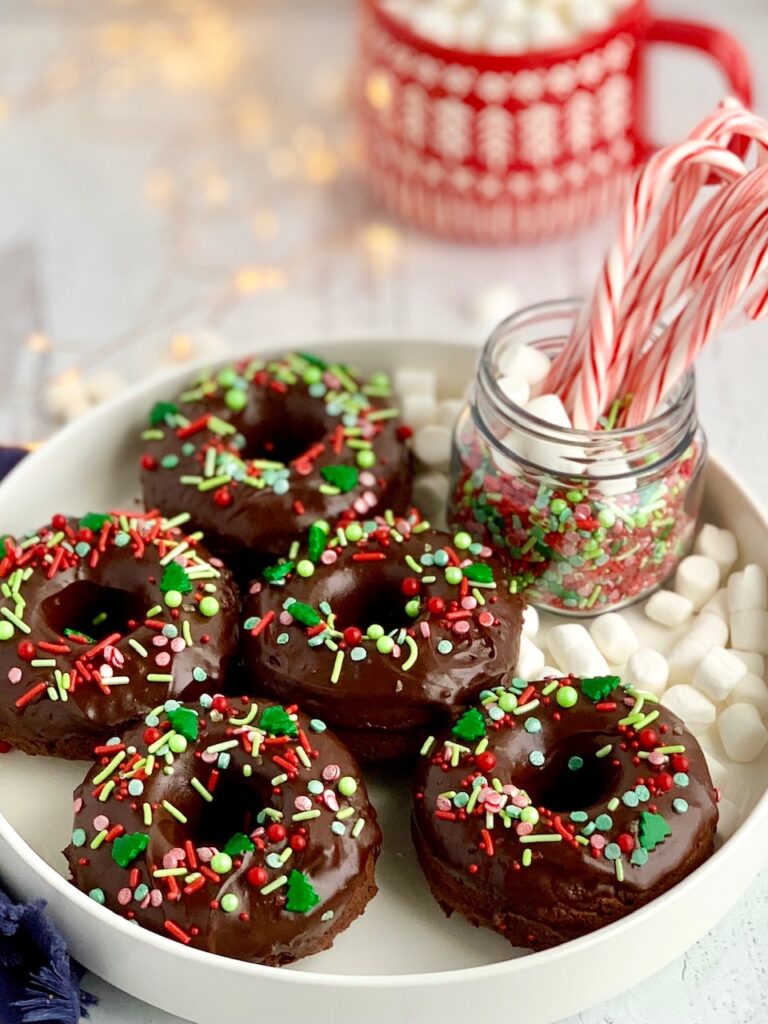 5 double chocolate donuts on a platter with chocolate ganache frosting topped with festive Christmas sprinkles next to a jar of candy canes. 