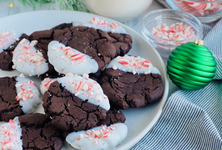 A white plate full of dark brown round cookies with half with half of the cookie dipped in melted white chocolate and candy cane pieces sprinkled on top.
