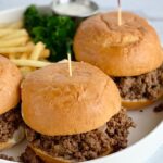 A white platter full of a meat mixture between hamburger buns with French fries.