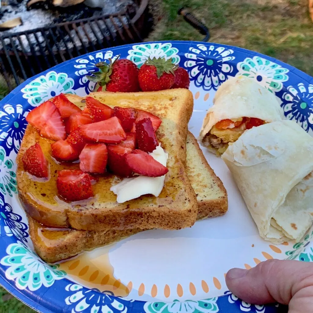 A plate with french toast, syrup, and sliced strawberries next to a breakfast burrito.