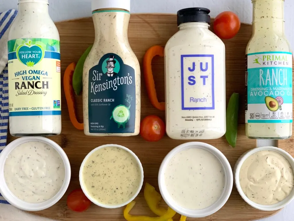 Four different dairy free bottles of ranch dressing laying down with a mini cup of dressing in front of each one to show what the dressing looks like.