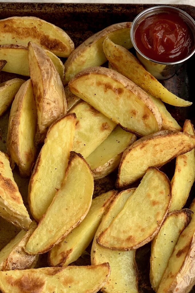 Baked potato wedges on a sheet pan with a container of ketchup
