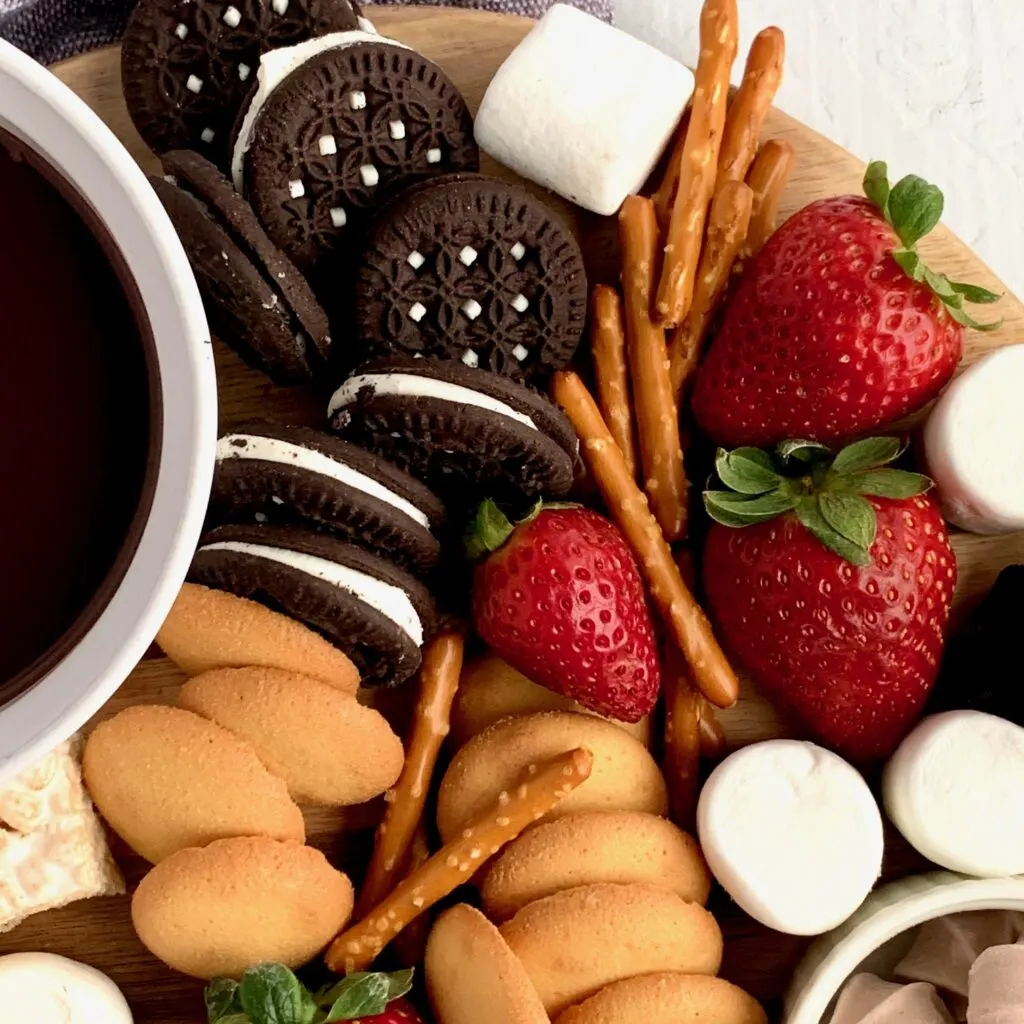 A dessert platter with strawberries, mini brownie bites, pretzel rods, oreo sandwich cookies, marshmallows, mini rice krispie treats and ganache dipping sauce.