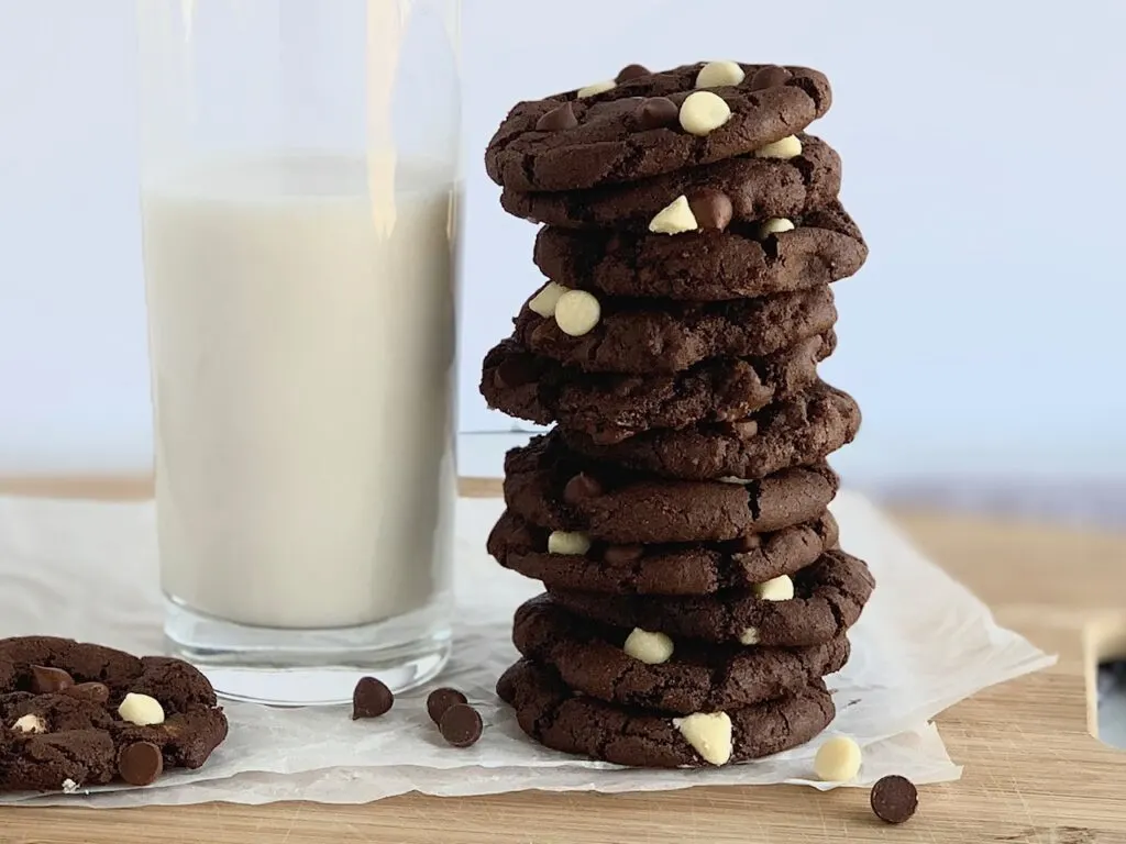 Several white chocolate chip cookies stacked next to a glass of almond milk.