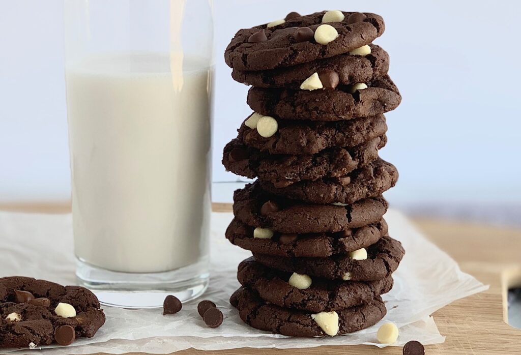 Several white chocolate chip cookies stacked next to a glass of almond milk.