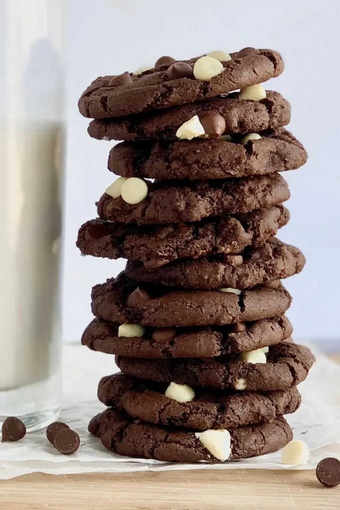 Several white chocolate chip cookies stacked next to glass of almond milk.