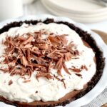 A white pie plate with a dark brown crust, a brown chocolate pudding, and topped with a white whipped topping. Last on top of that is small pieces of hard chocolate.