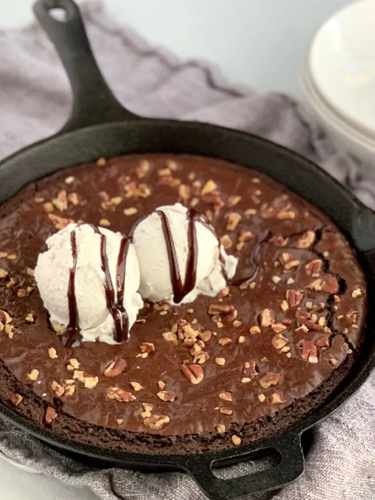 Baked brownie in a cast iron pan with walnut, ice cream, and chocolate syrup on top.