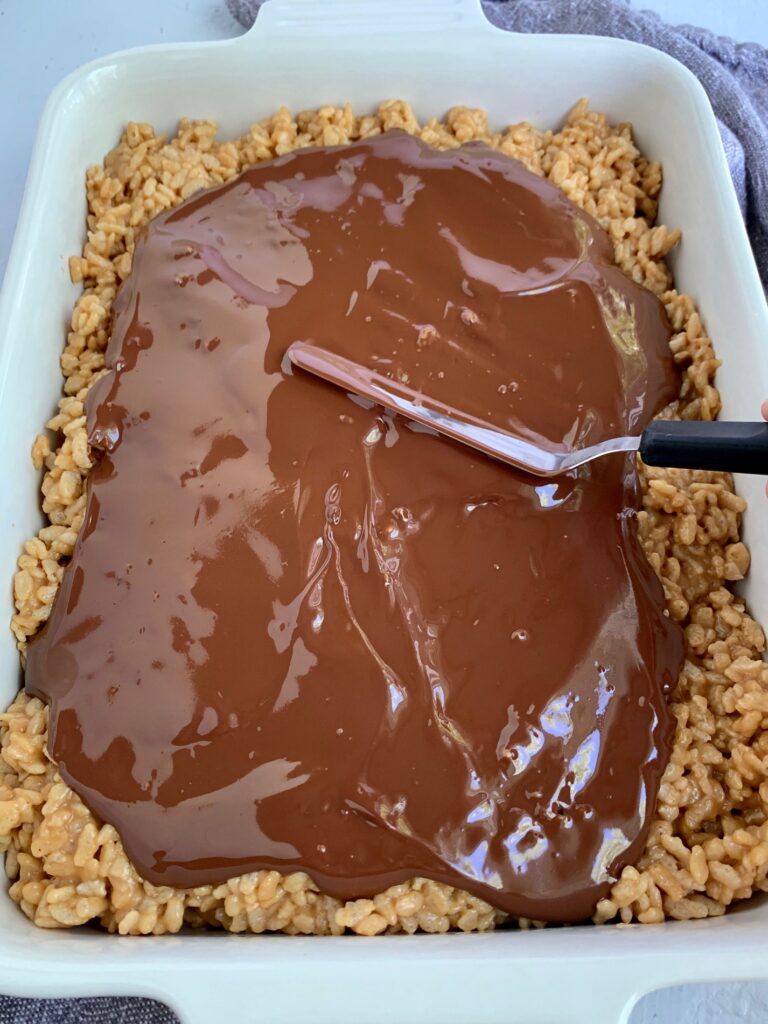 An angle icing spatula spreading melted chocolate on the peanut butter cereal mixture.
