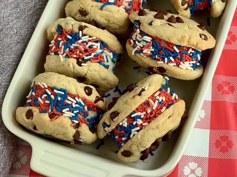 In a white serving dish are chocolate chip cookies with ice cream and red, white, and blue sprinkles on them.