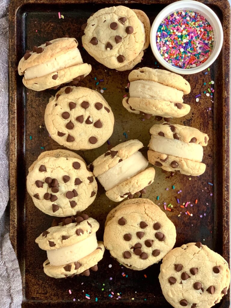 Ice cream sandwiched between two chocolate chip cookies on a large cookie pan with sprinkles on it.