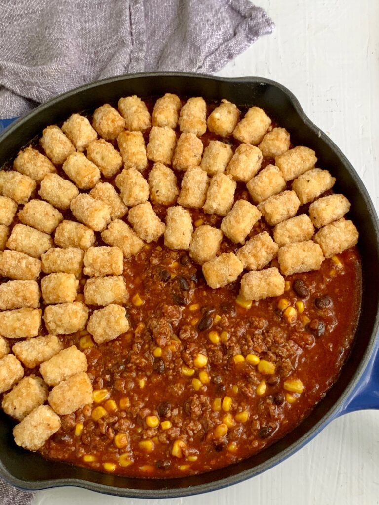 Putting tater tots on top of a taco mixture of ground beef, corn, black beans, and sauce mixture. 