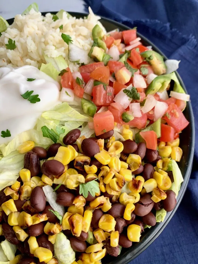 A bowl filled with iceberg lettuce, chicken, corn, black beans, cilantro lime rice, pico de gallo, and a dollop of sour cream.