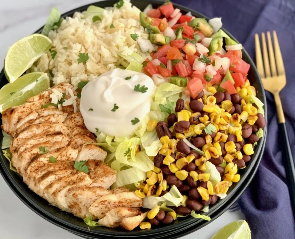 A bowl filled with iceberg lettuce, chicken, corn, black beans, cilantro lime rice, pico de gallo, and a dollop of sour cream.