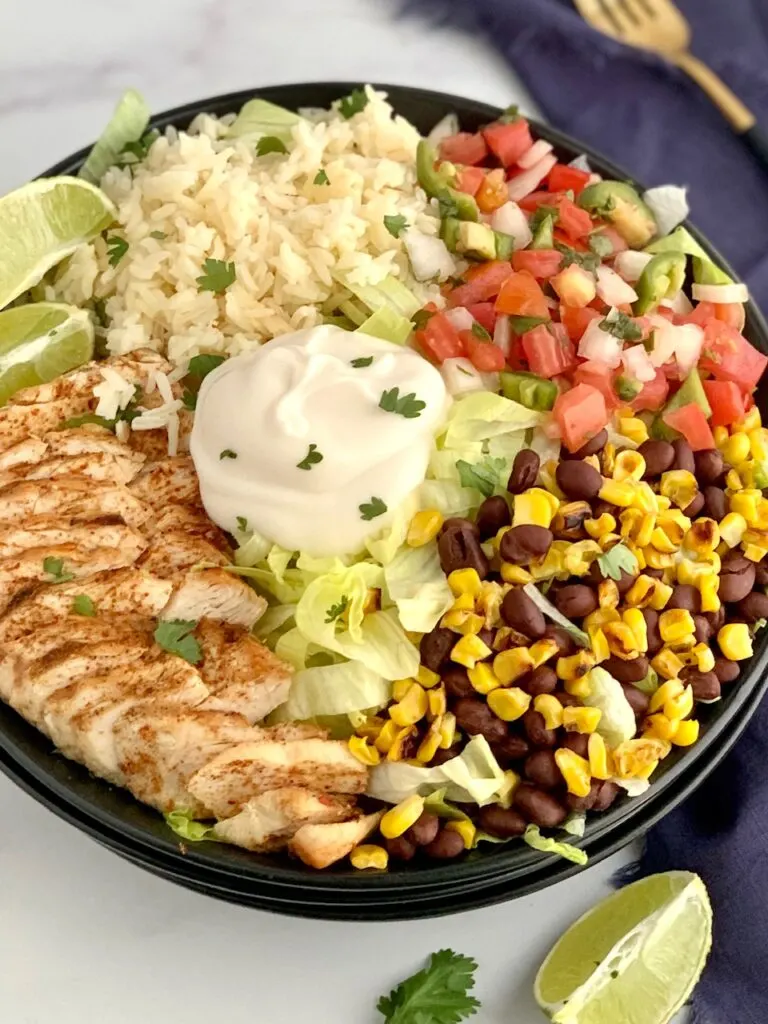 A bowl filled with iceberg lettuce, chicken, corn, black beans, cilantro lime rice, pico de gallo, and a dollop of sour cream.