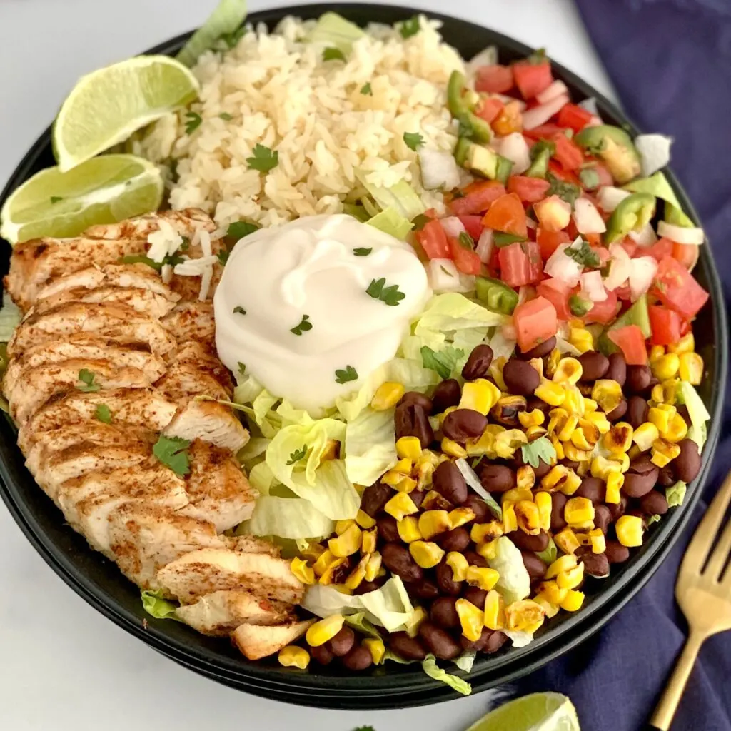 A bowl filled with iceberg lettuce, chicken, corn, black beans, cilantro lime rice, pico de gallo, and a dollop of sour cream.