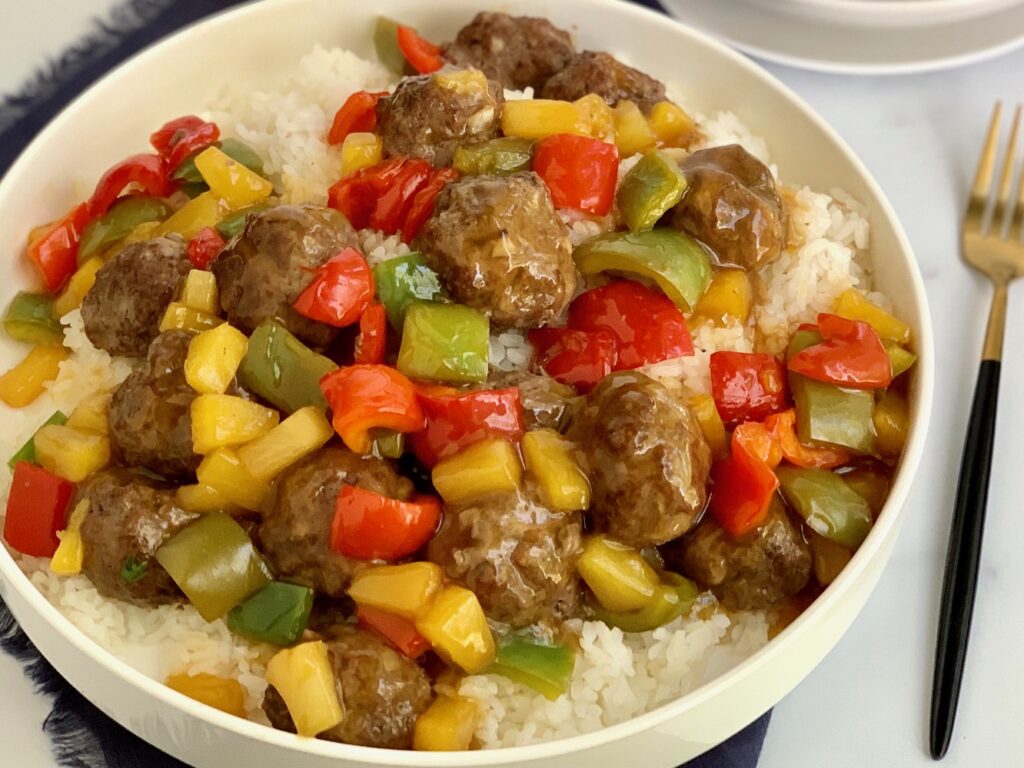 A bowl of white rice with meatballs, pineapple chunks, and bell peppers. 