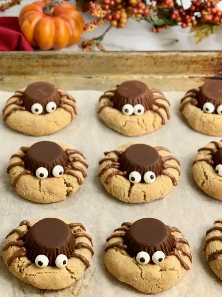 a baking sheet with peanut butter cookies topped with an upside down peanut butter cup, four frosting legs on each side and 2 googly eyes to look like a spider on  cookie