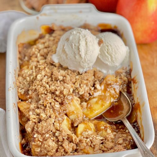 A white baking dish filled with thin apple slices topped with a buttery, brown sugar, and oat topping. On top of that is 2 scoops of dairy free ice cream starting to melt.