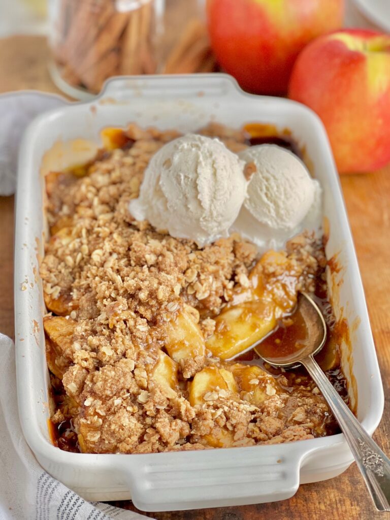 A white baking dish filled with thin apple slices topped with a buttery, brown sugar, and oat topping. On top of that is 2 scoops of dairy free ice cream starting to melt.