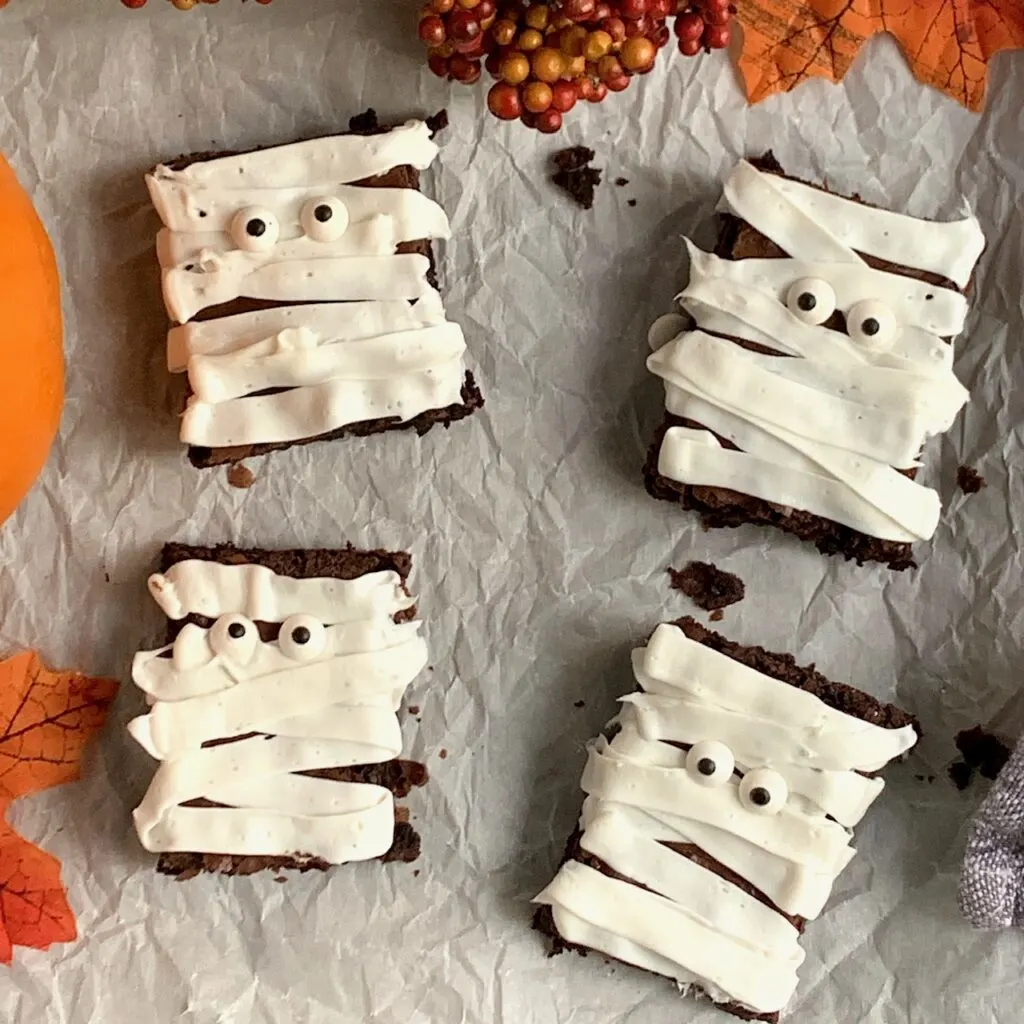 4 rectangular brownie's with white frosting draped across and google eyes to look like mummies