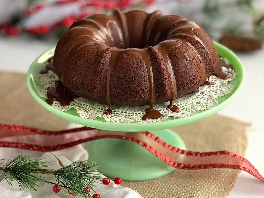 A chocolate bundt cake on a green cake plate with a chocolate ganache frosting running down the sides. 